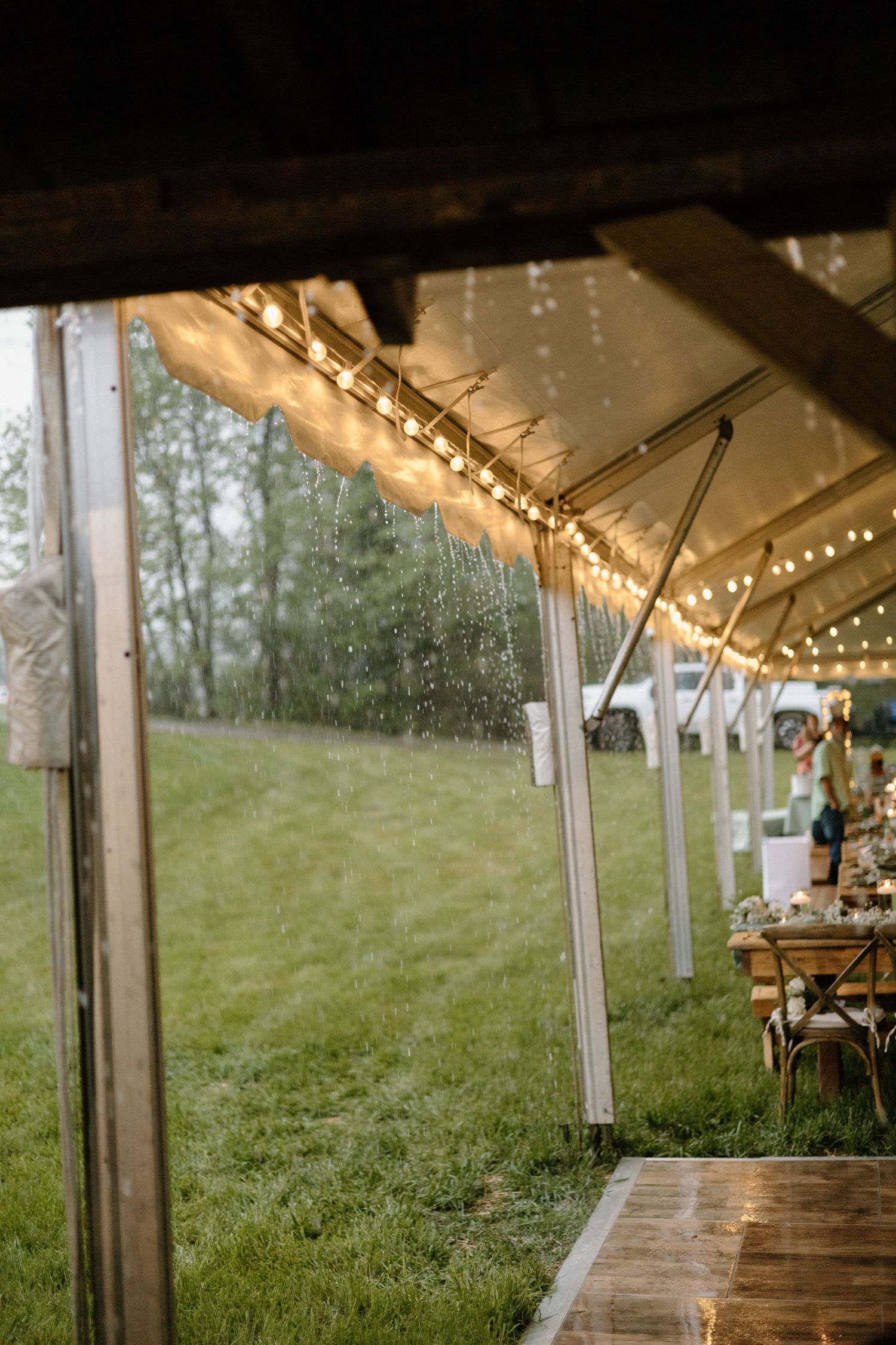 A wedding tent decorated with string lights and wooden tables set up on a grassy hillside during golden hour.