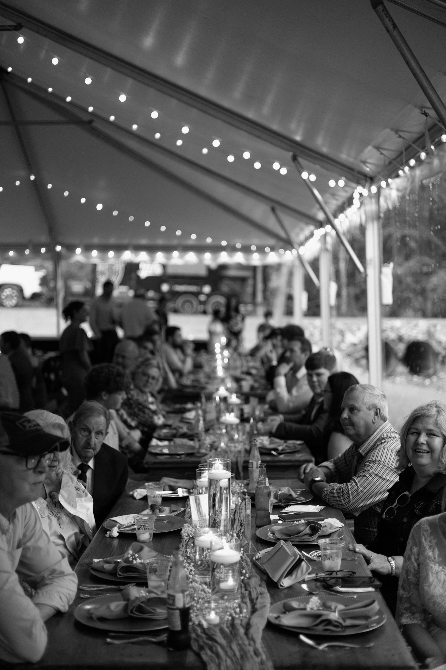 Black and white photo series of a wedding reception dinner under a tent with string lights and guests seated at long tables.