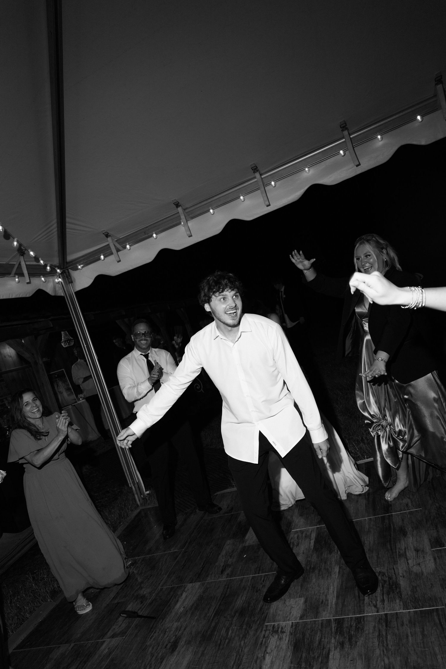 Black and white photo of people dancing under a tent at a party with string lights overhead on a wooden dance floor.