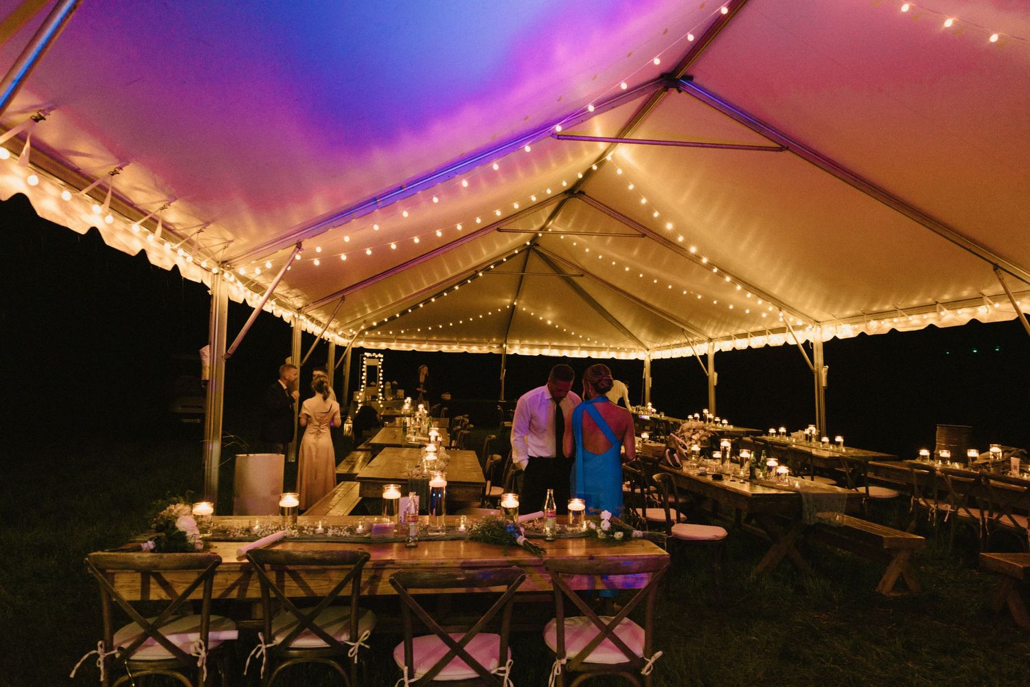 String lights illuminate a white wedding tent at dusk creating a romantic outdoor reception atmosphere.