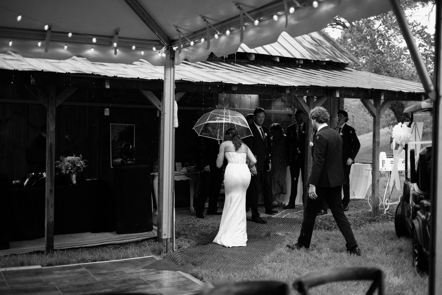 A black and white panoramic photo shows a wedding reception under a tent with string lights and guests mingling.