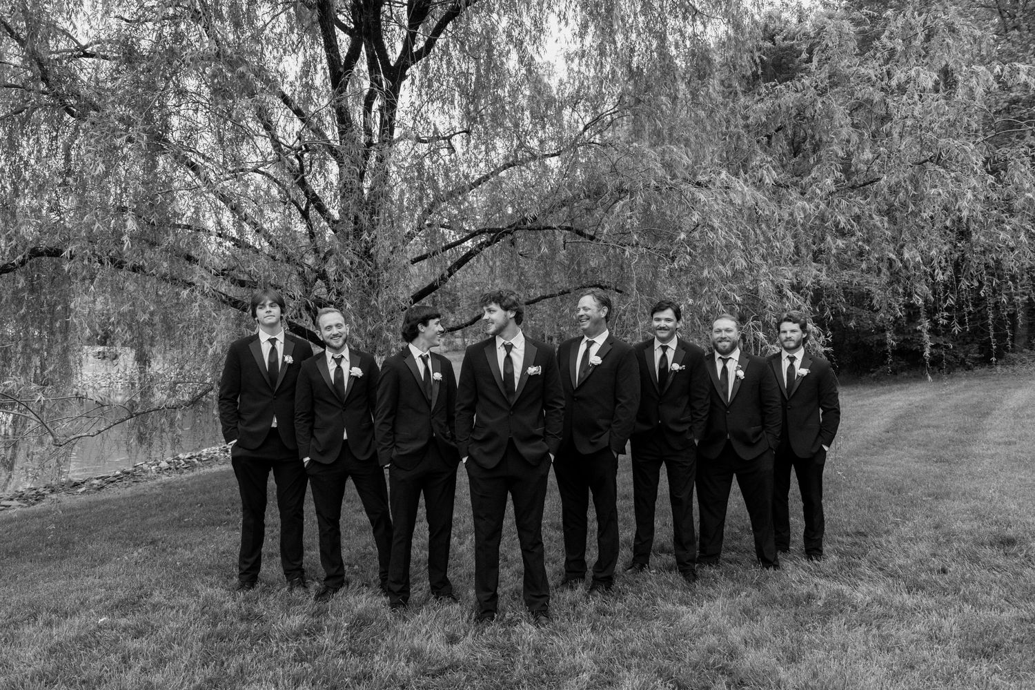 A group of groomsmen in black suits stand together outdoors under a willow tree in black and white photography.