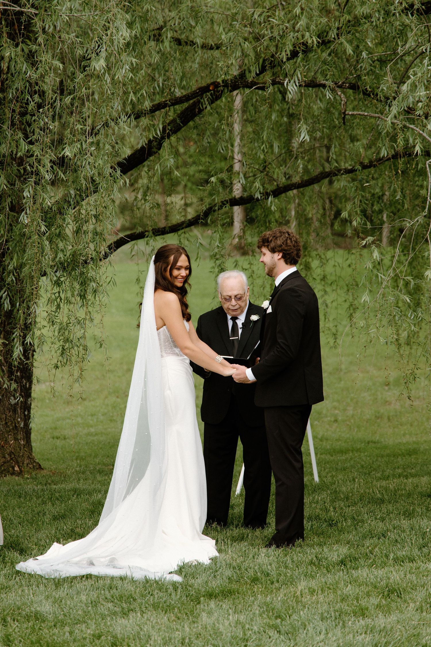 An outdoor wedding ceremony takes place under drooping willow trees on a lush green lawn with two people facing an officiant.