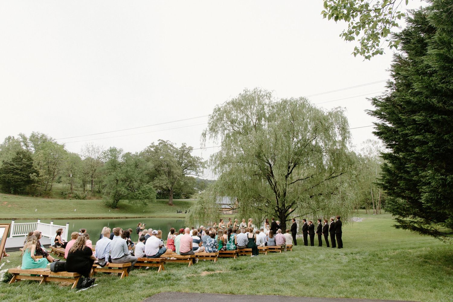 An outdoor wedding ceremony setup with wooden chairs on a grassy lawn surrounded by tall trees in a park-like setting.
