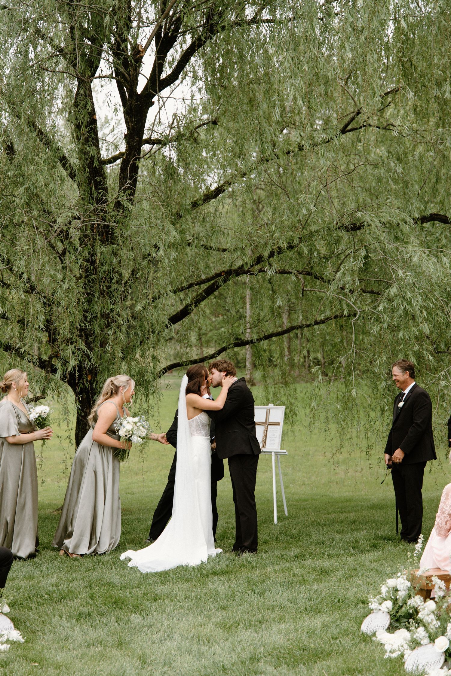 An outdoor wedding ceremony under a weeping willow tree with bridesmaids in gray dresses standing on lawn.
