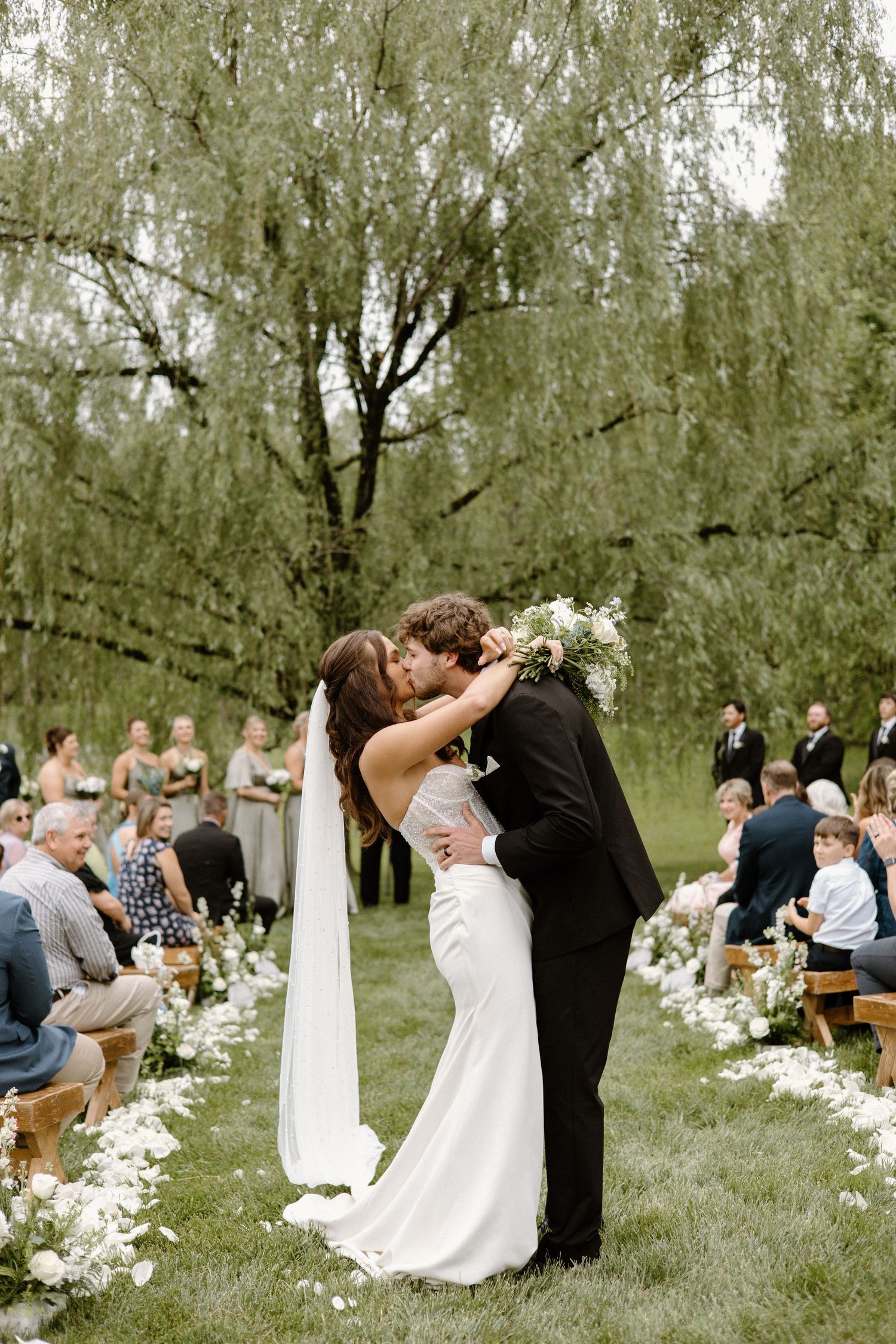 A couple shares their first kiss at an outdoor garden wedding ceremony with guests seated on wooden benches.