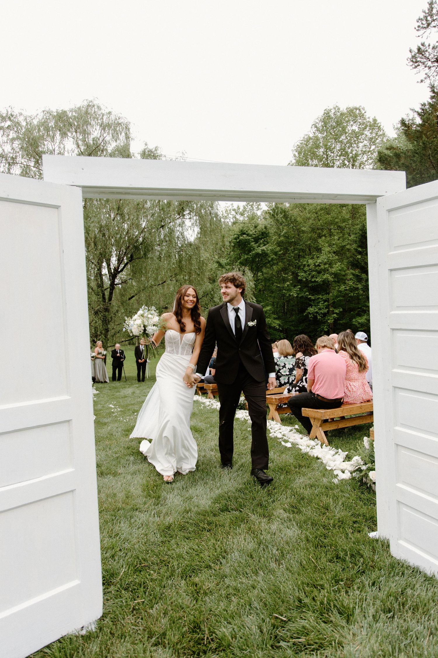 Outdoor wedding ceremony set up with white doors and wooden bench in a grassy garden setting.