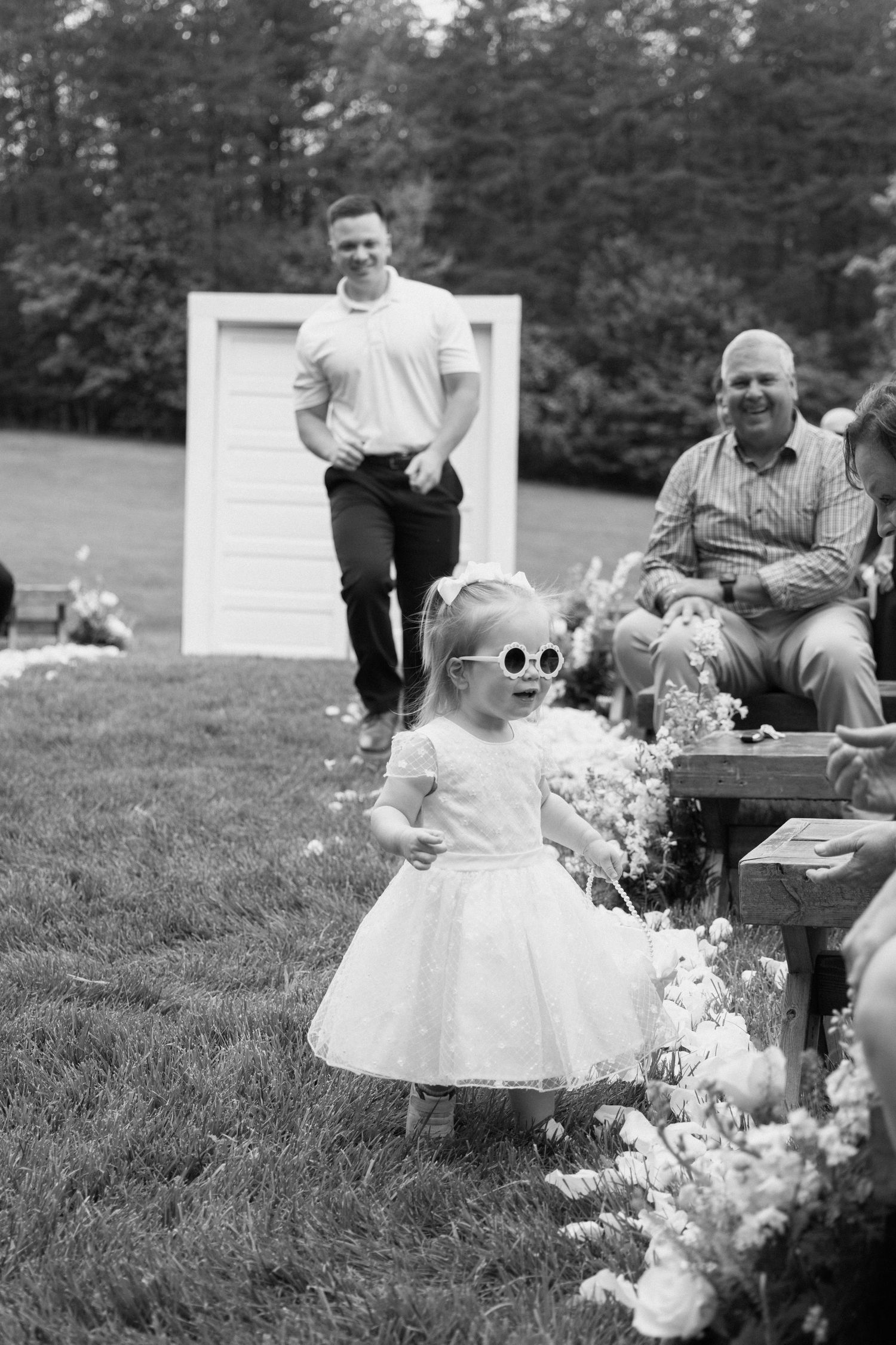 Small child in a white dress plays in a grassy field during an outdoor family gathering with picnic tables.