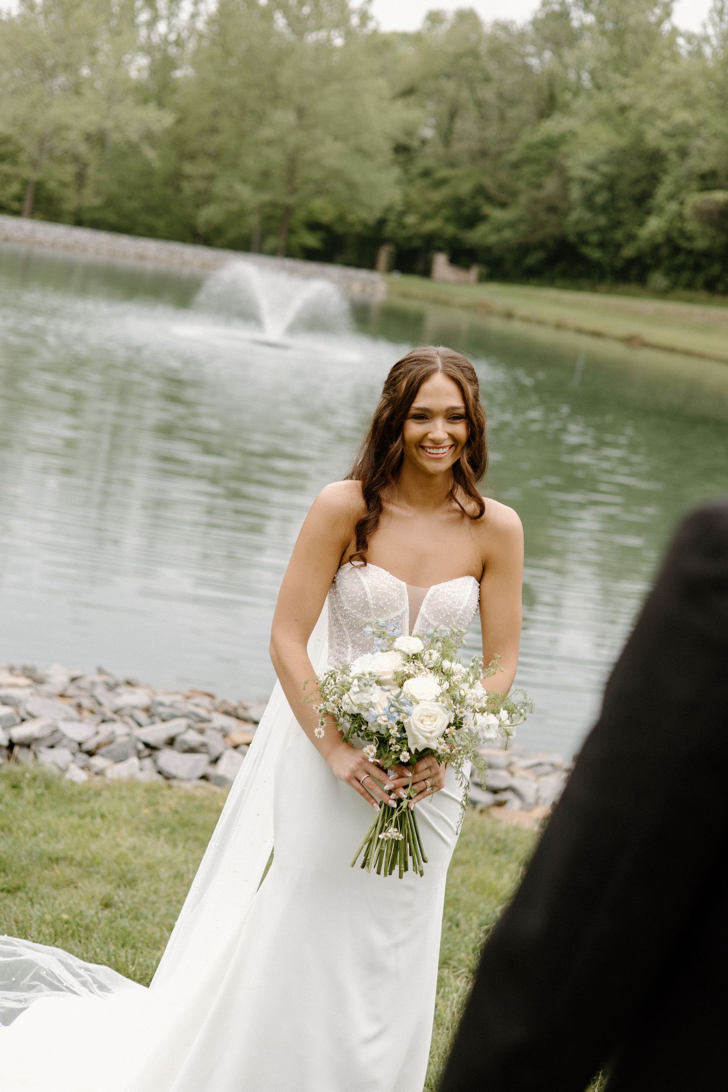 Beautiful wedding scene by a pond with fountain showing a person in white gown holding elegant white floral bouquet.