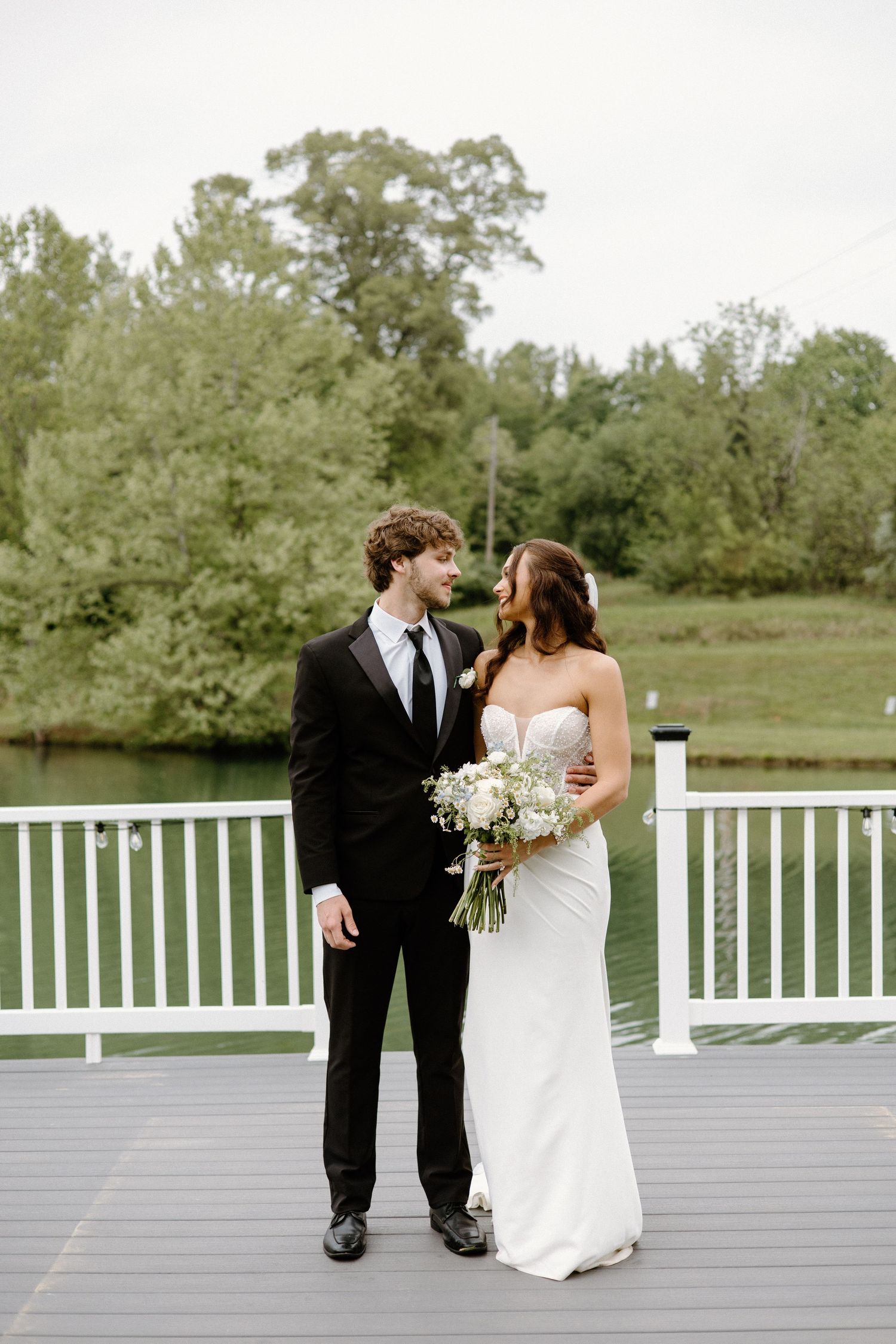 Newlywed couple stands together on a gray deck with white railing overlooking lush green grounds on their wedding day.