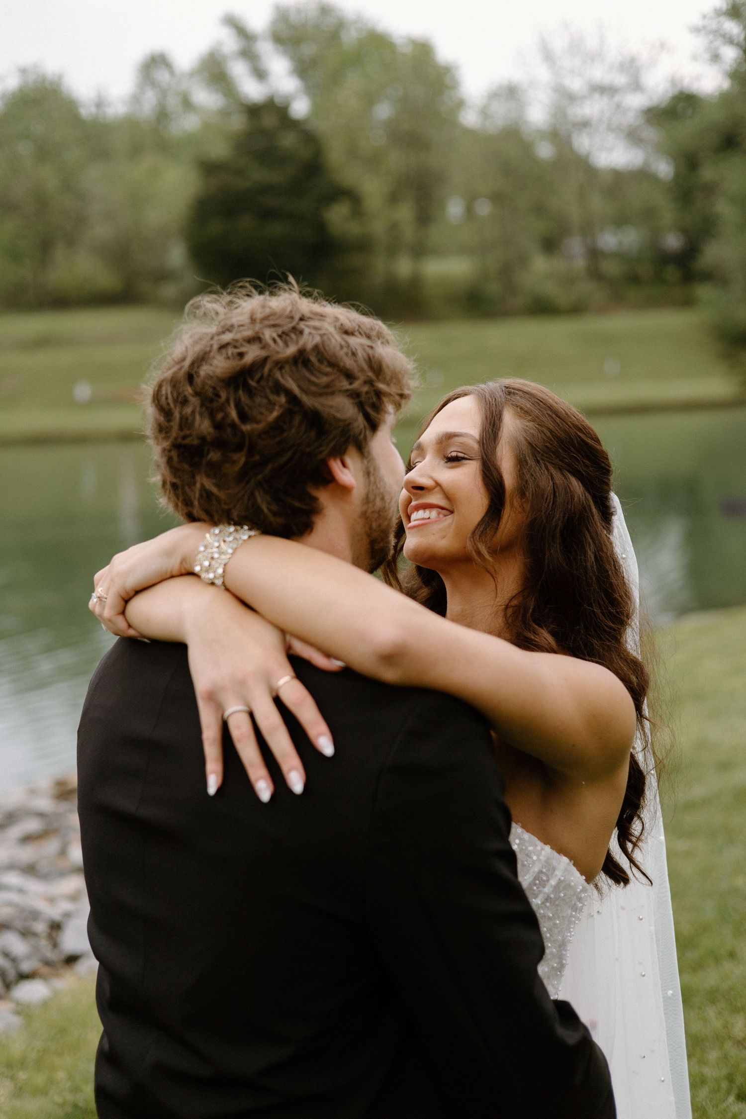 A couple embraces outdoors by a serene lake, with the bride wearing a white dress and groom in a black suit.