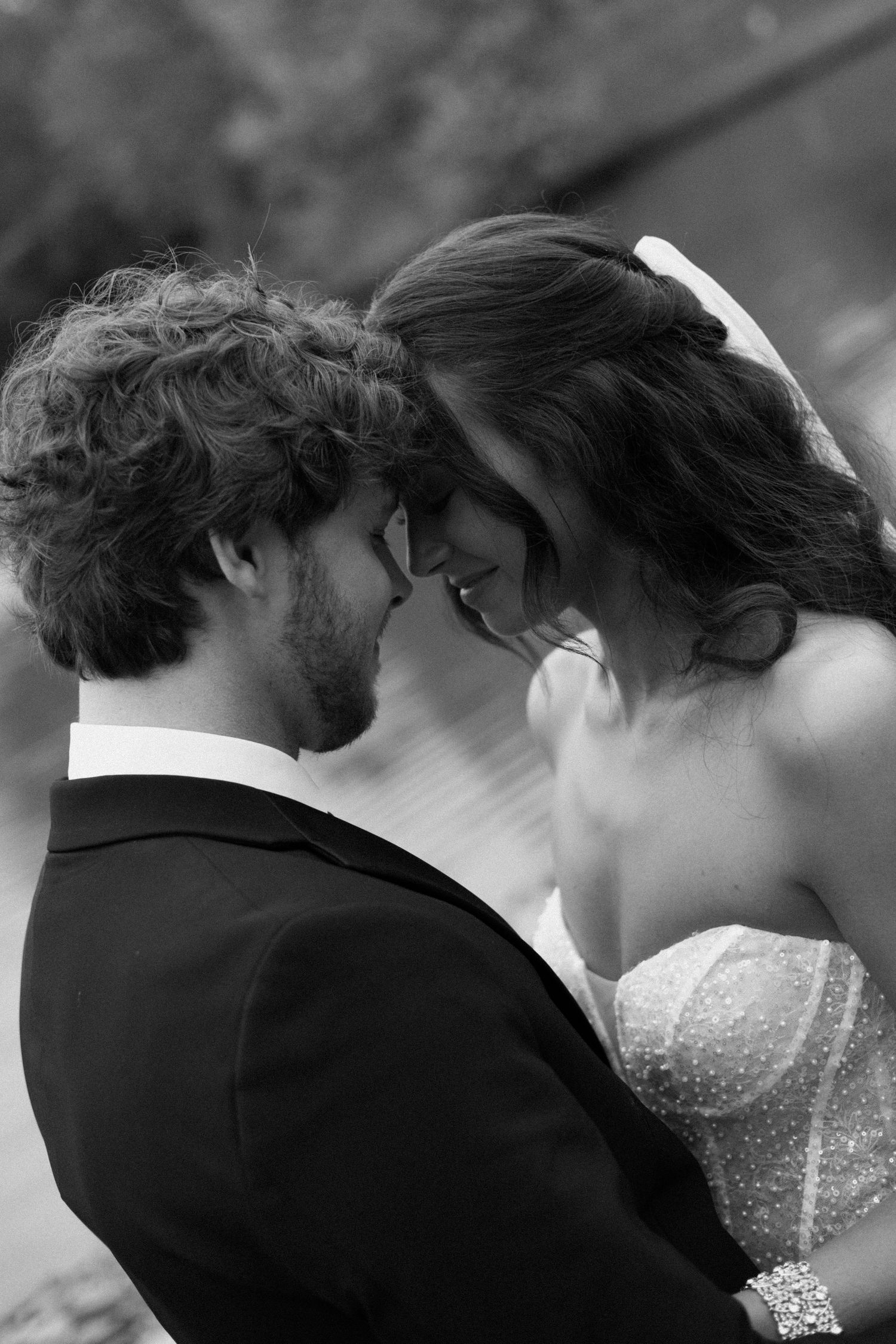 Black and white romantic wedding photo showing bride and groom in intimate forehead-touching pose outdoors.