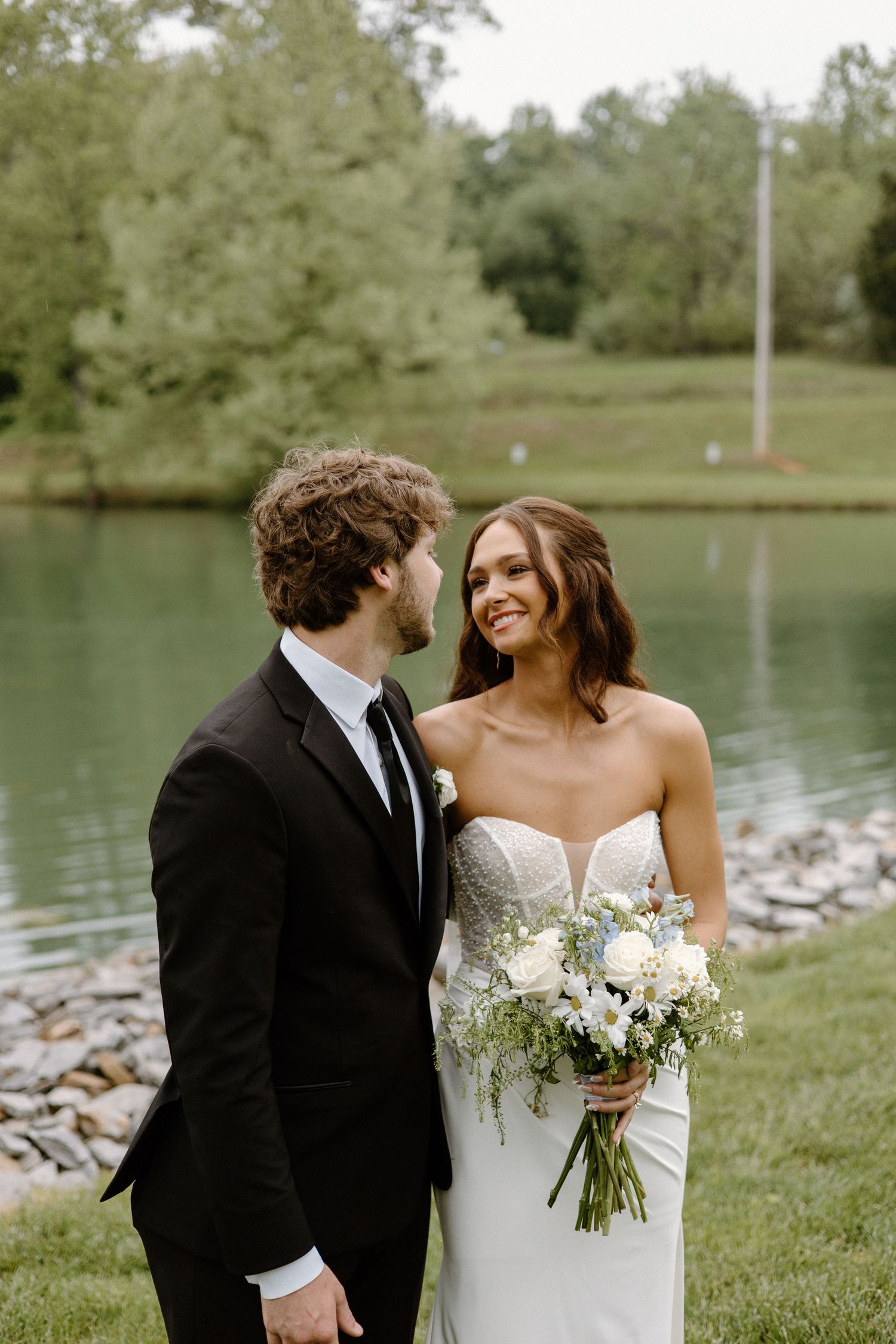 Newlywed couple shares intimate moments by a serene lake in a picturesque outdoor setting.