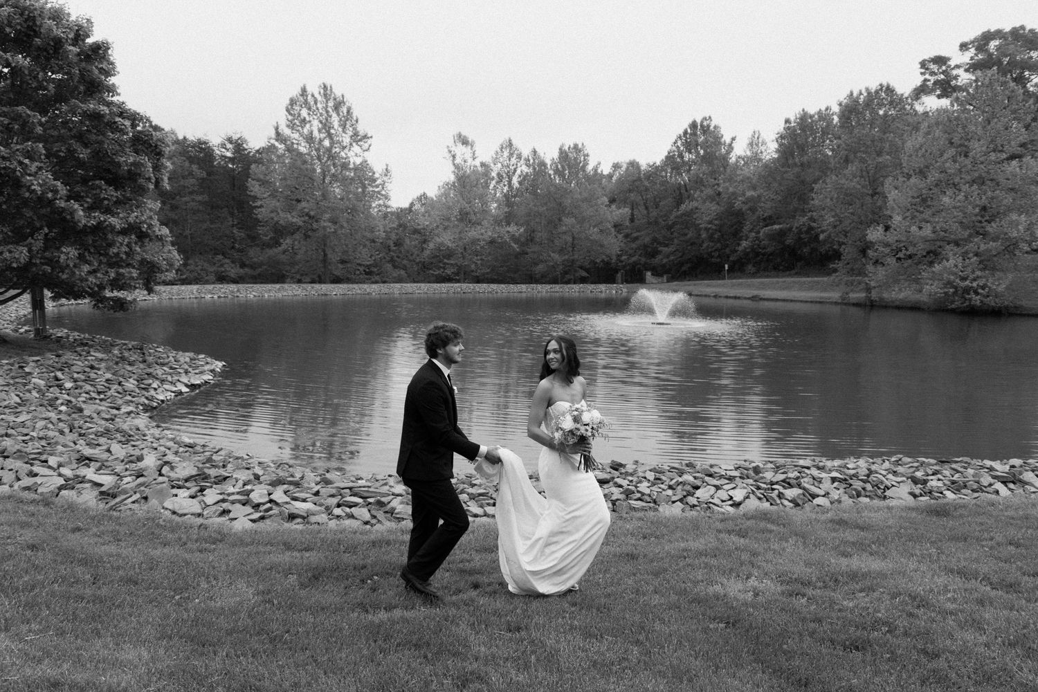 A couple walks along a lakeside path with lush trees and landscaping on the grounds of an estate.