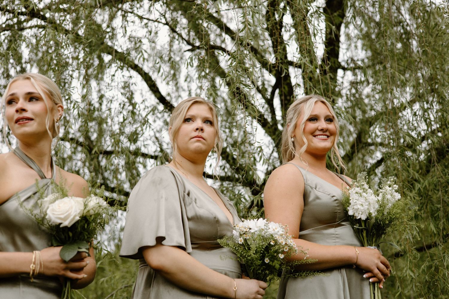 A group of people outdoors smile and laugh together while holding champagne glasses near trees.