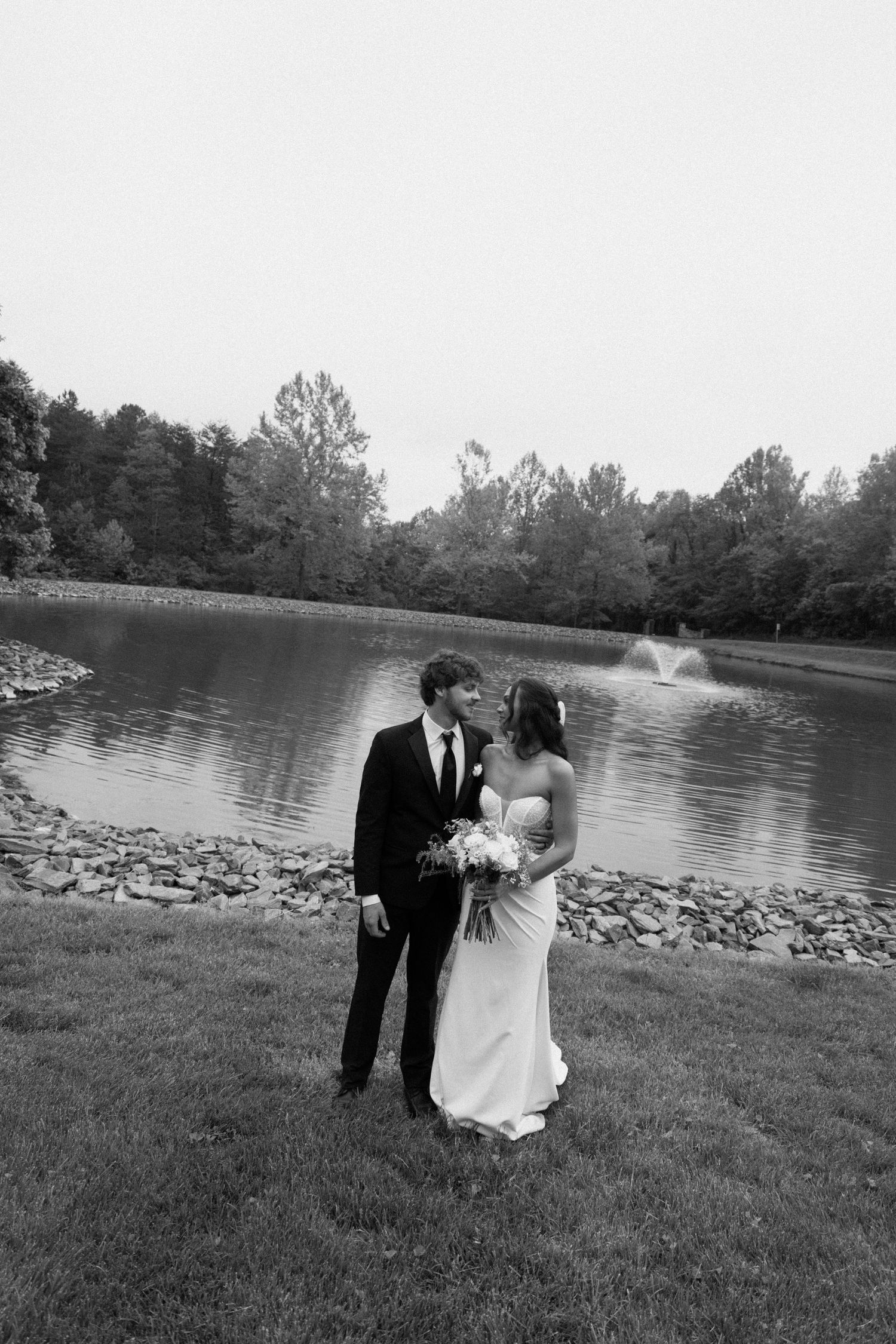 A black and white photo of newlyweds sharing an intimate moment beside a scenic lake with water fountain and trees.