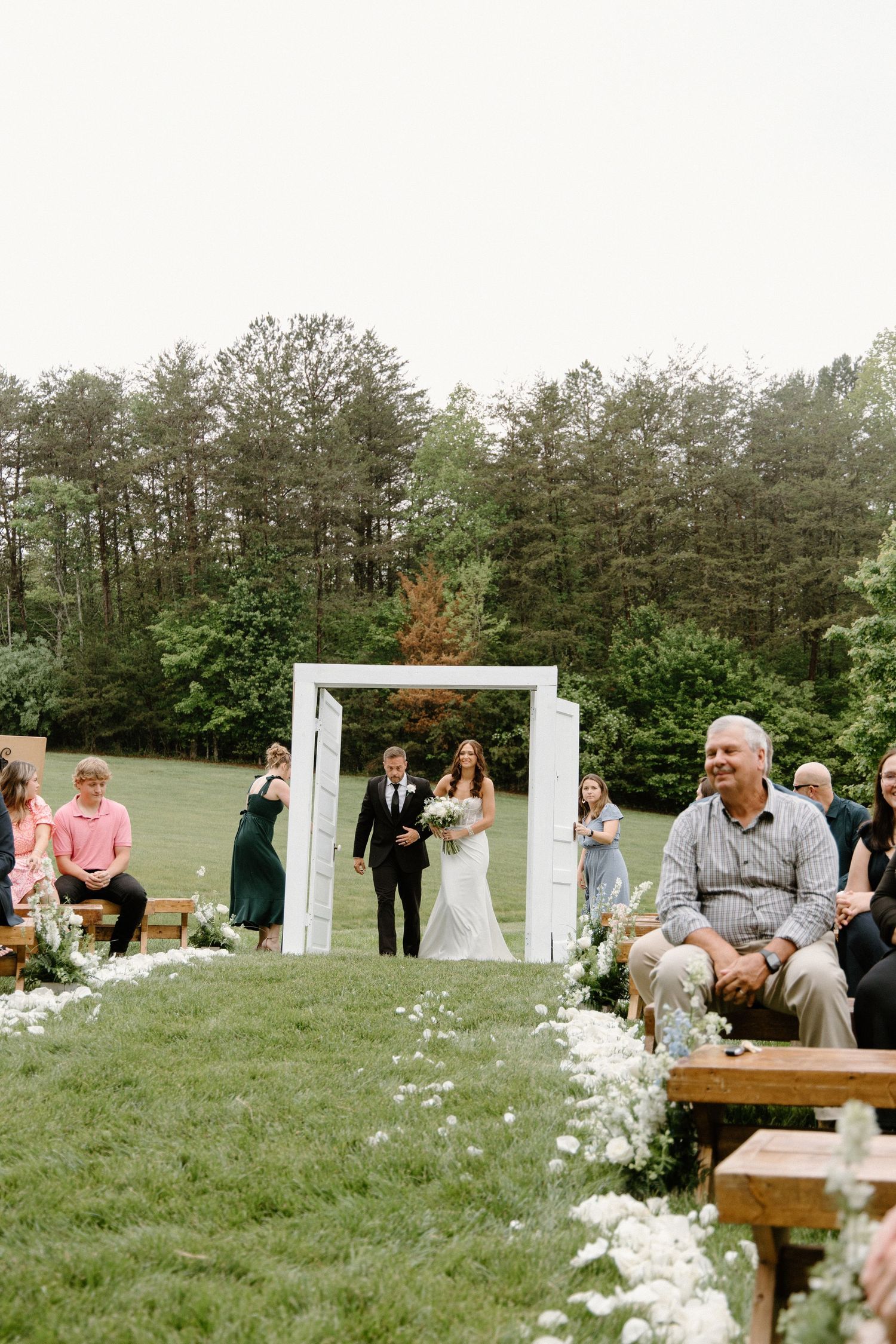 Outdoor wedding ceremony on grassy lawn with white arch and wooden benches decorated with flower petals along aisle.