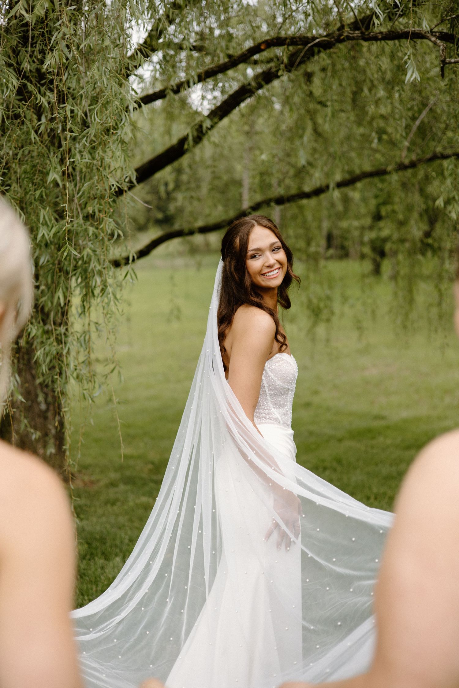 A bride in a flowing white veil twirls playfully under a willow tree in a garden setting.