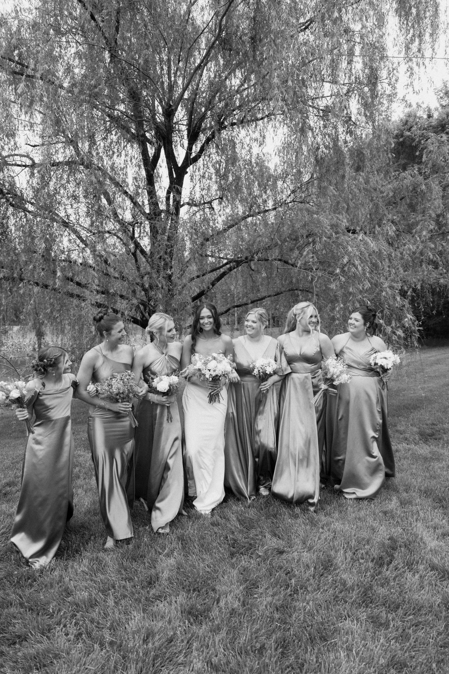 Black and white photo of a bridal party in elegant floor-length gowns gathered outdoors under a weeping willow tree.