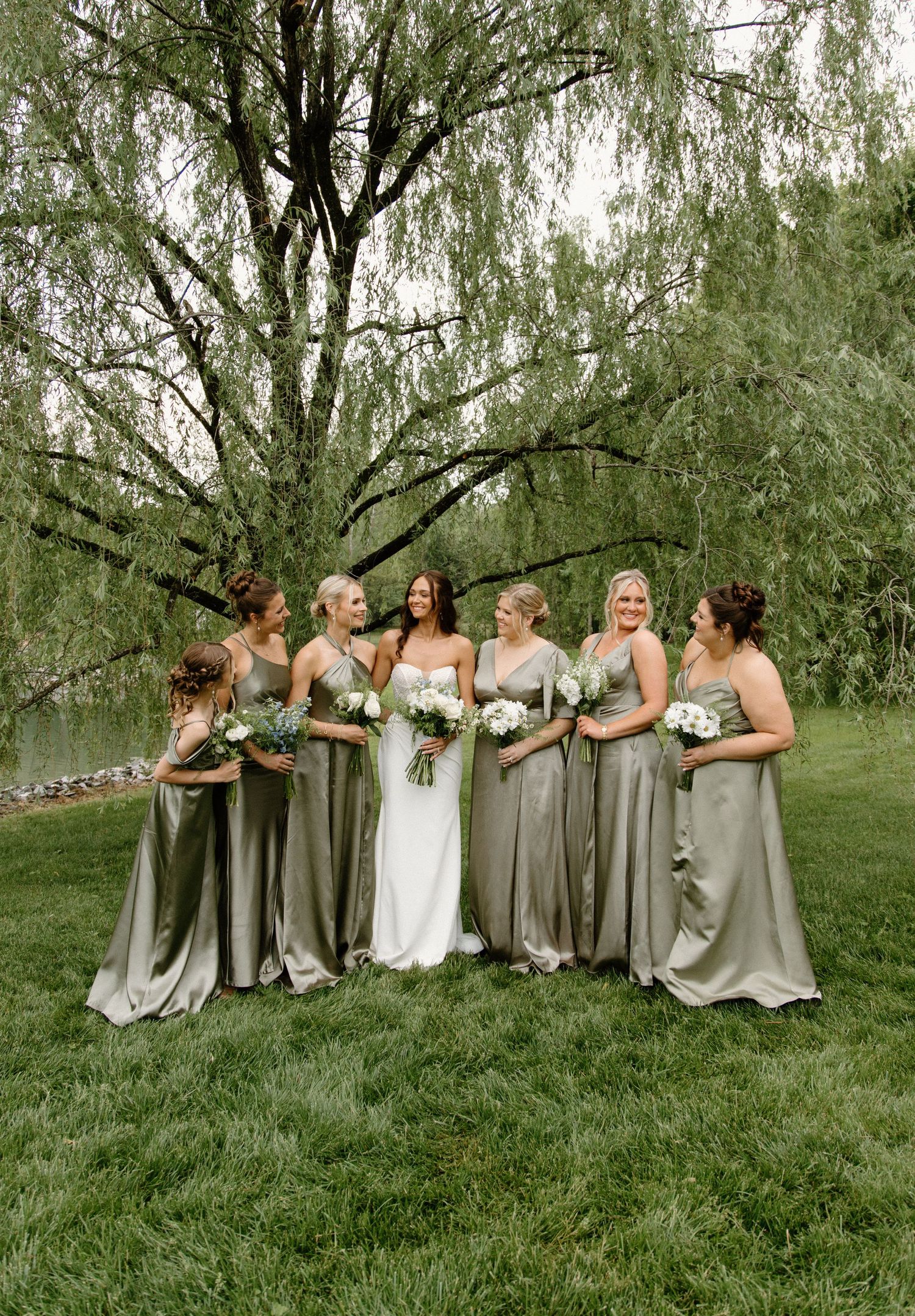 Wedding party poses on green lawn beneath graceful willow tree wearing elegant silver bridesmaid dresses.