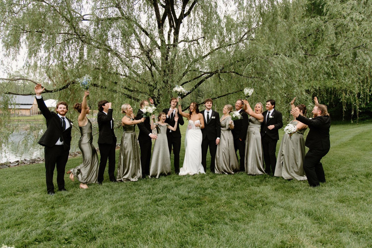 Wedding party in formal attire celebrates outdoors with joyful poses under a weeping willow tree on green lawn.