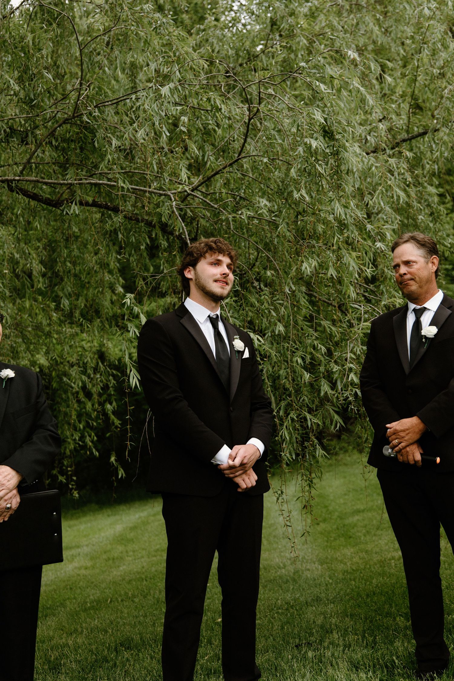 A group in black suits poses outdoors with a lush background of green willow trees.