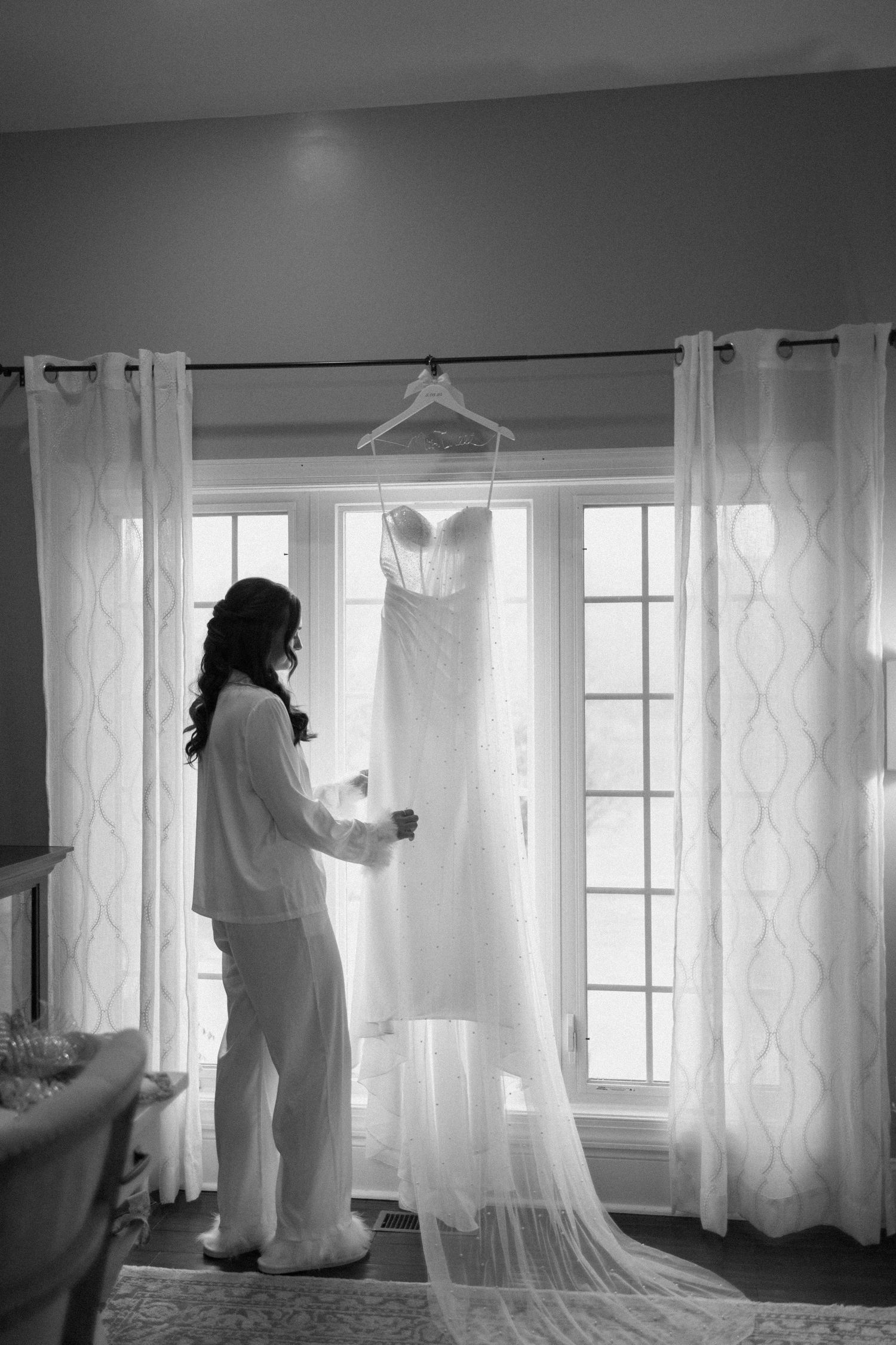 Black and white photo of a bride's wedding dress hanging from a curtain rod while someone admires it by a window.