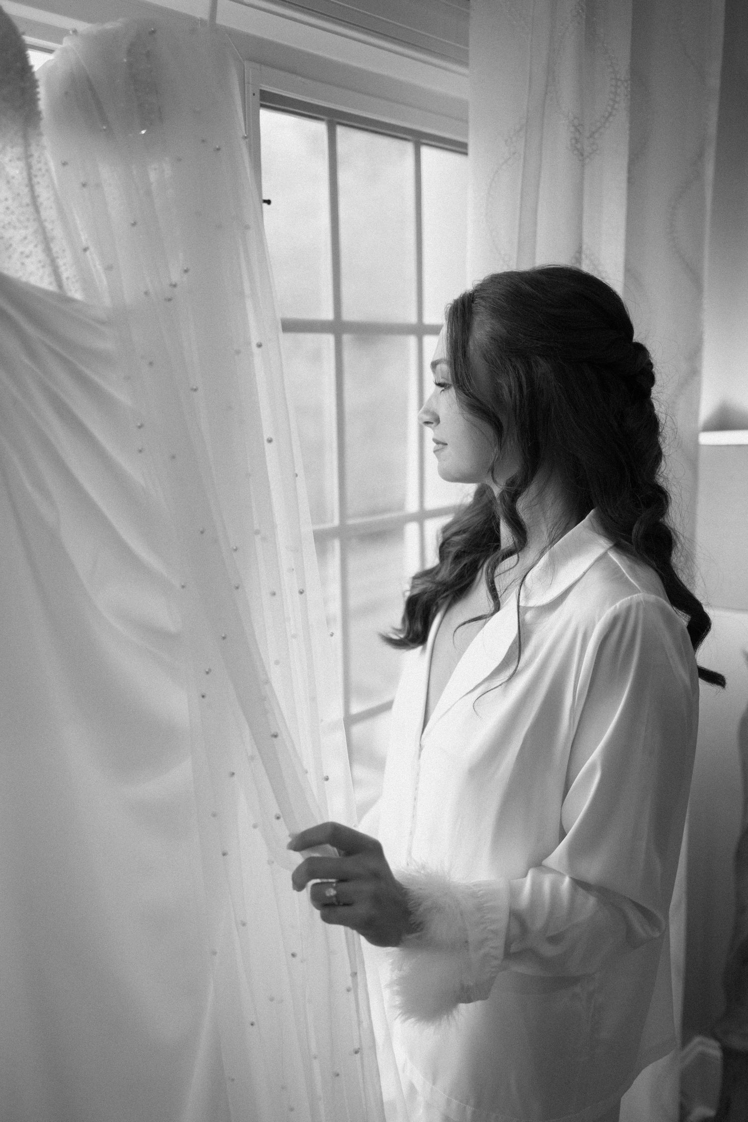 A woman in white robe stands contemplatively by window near wedding dress in black and white.