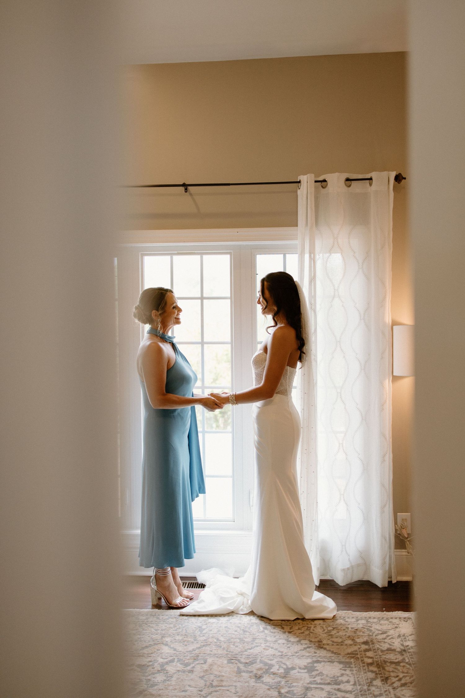 Two figures stand in silhouette by a sunlit window, one wearing a light blue dress and one in a fitted white gown.