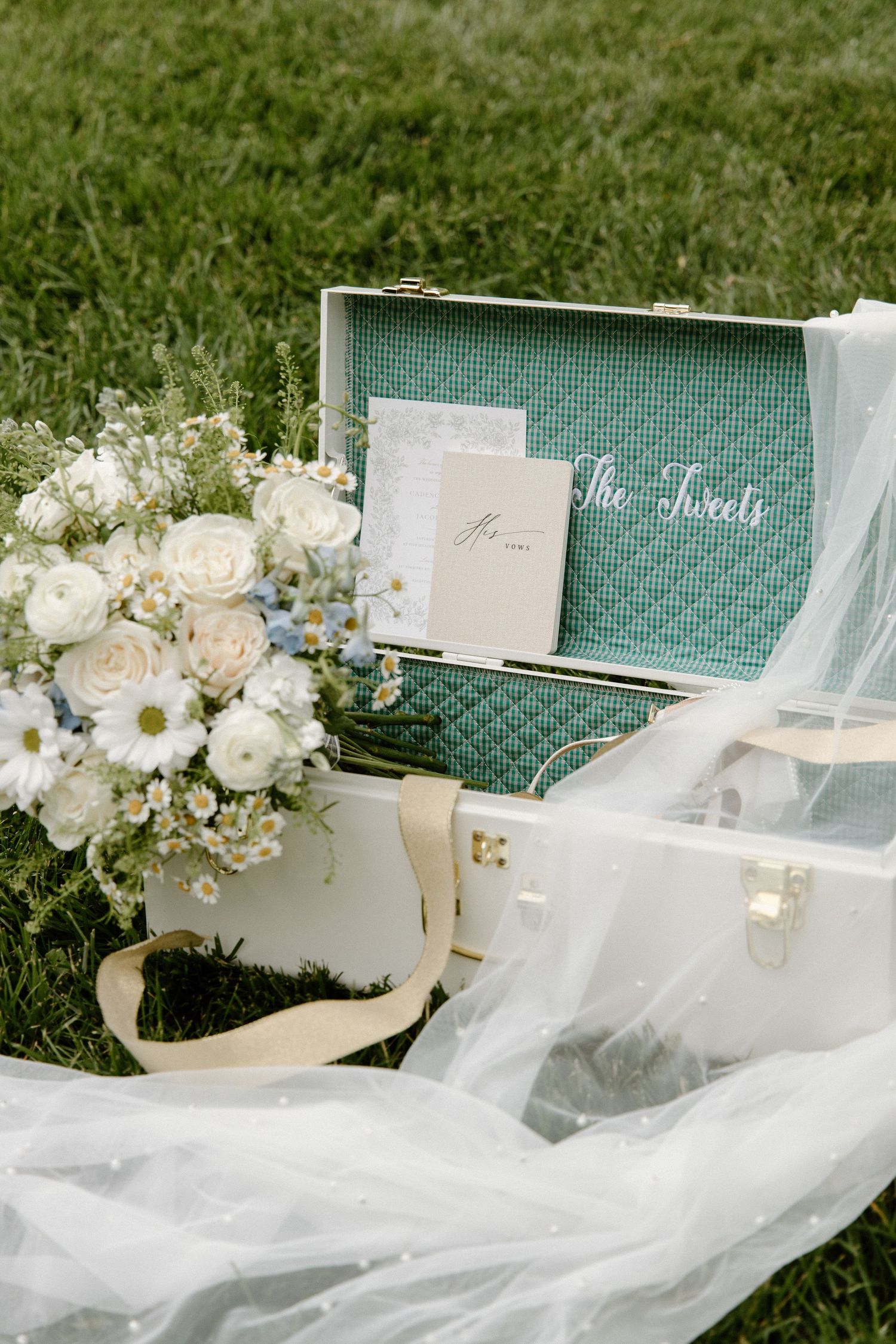 White wedding flowers and gauzy veil arranged with vintage turquoise suitcase on grass.