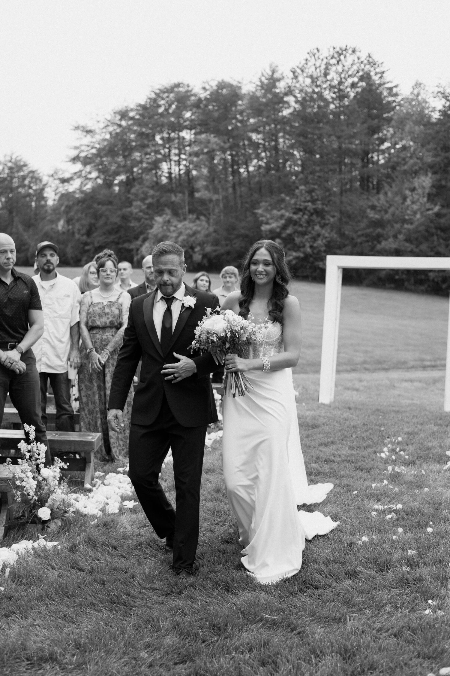 A couple walks down an outdoor aisle at their wedding ceremony with guests and trees in the background.