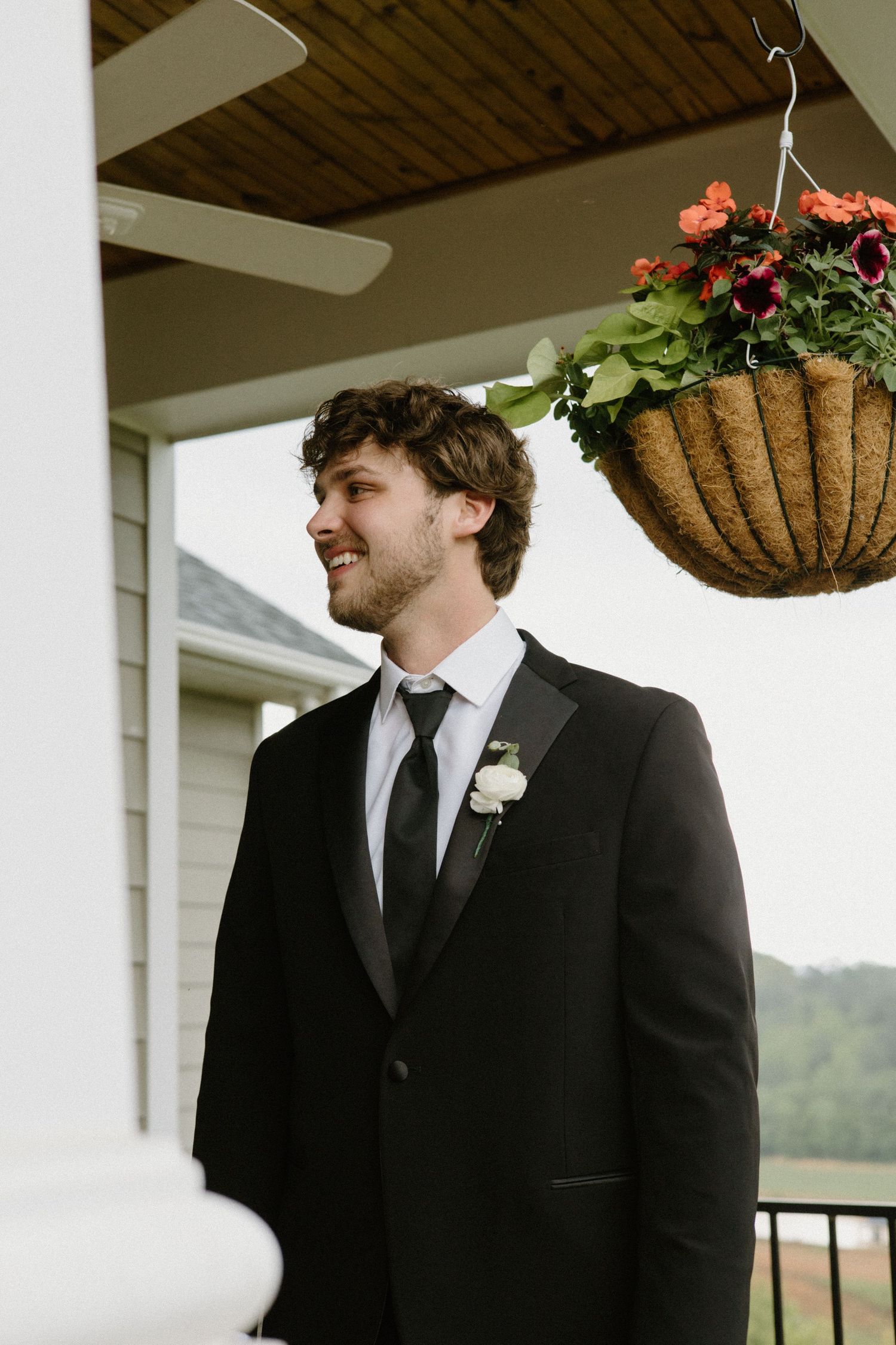 Groomsmen in black suits and ties stand on a white porch with hanging flower baskets.