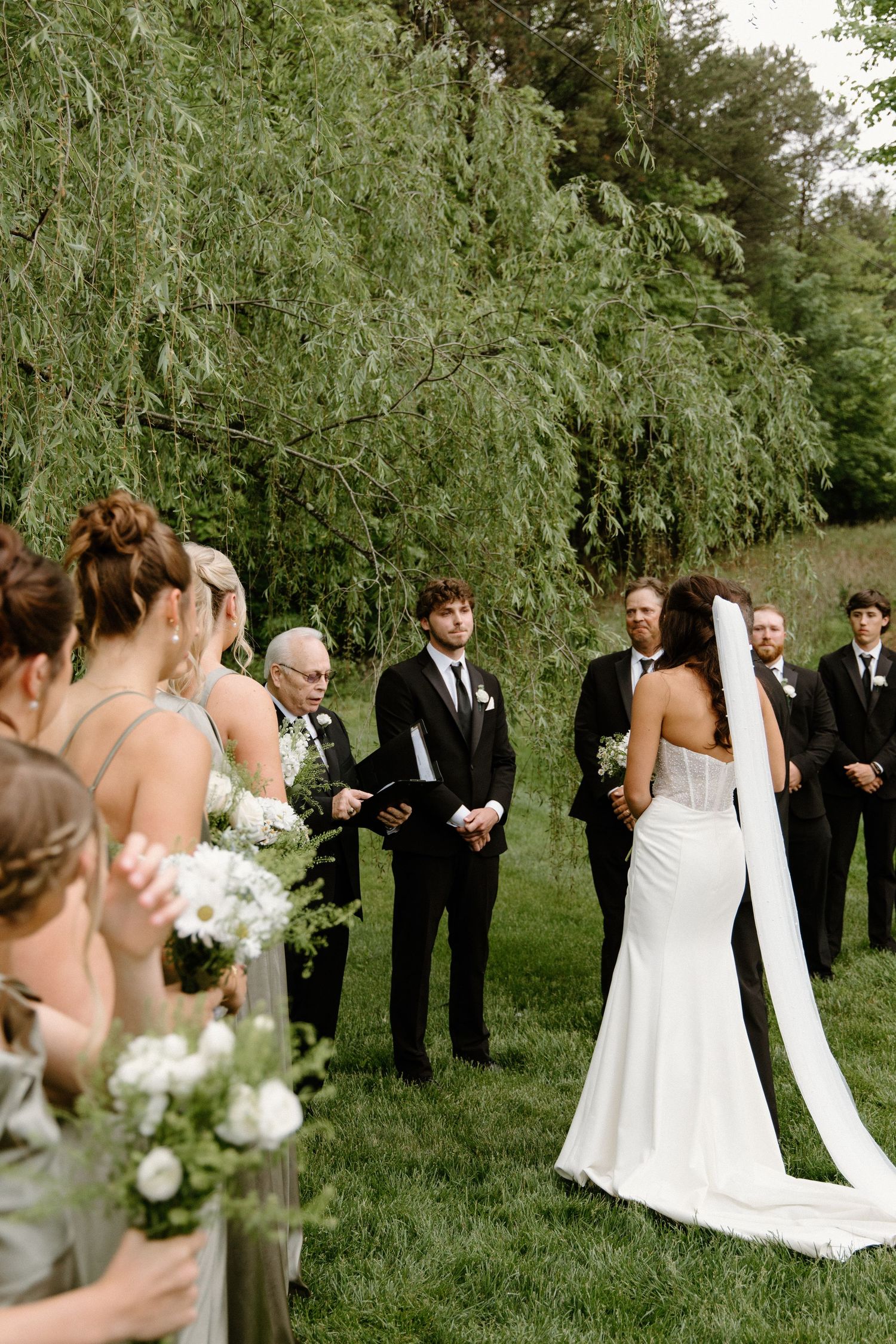 Wedding party stands under drooping willow trees on a lush green lawn holding white floral bouquets.