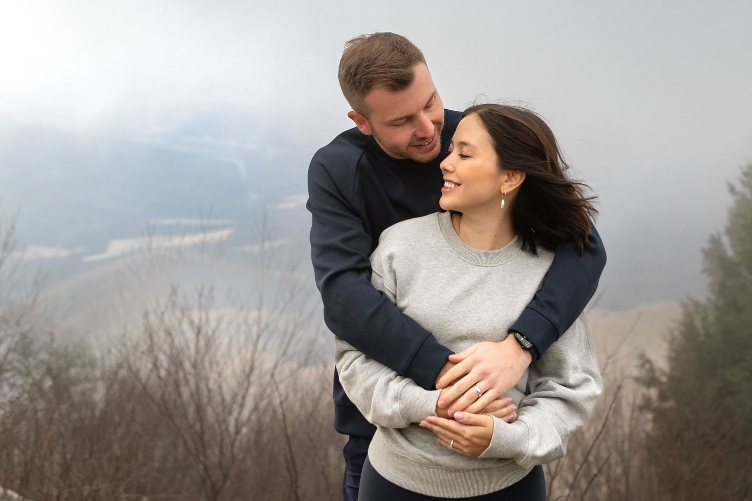 A couple embraces while standing on a foggy hillside in winter wearing cozy sweaters.