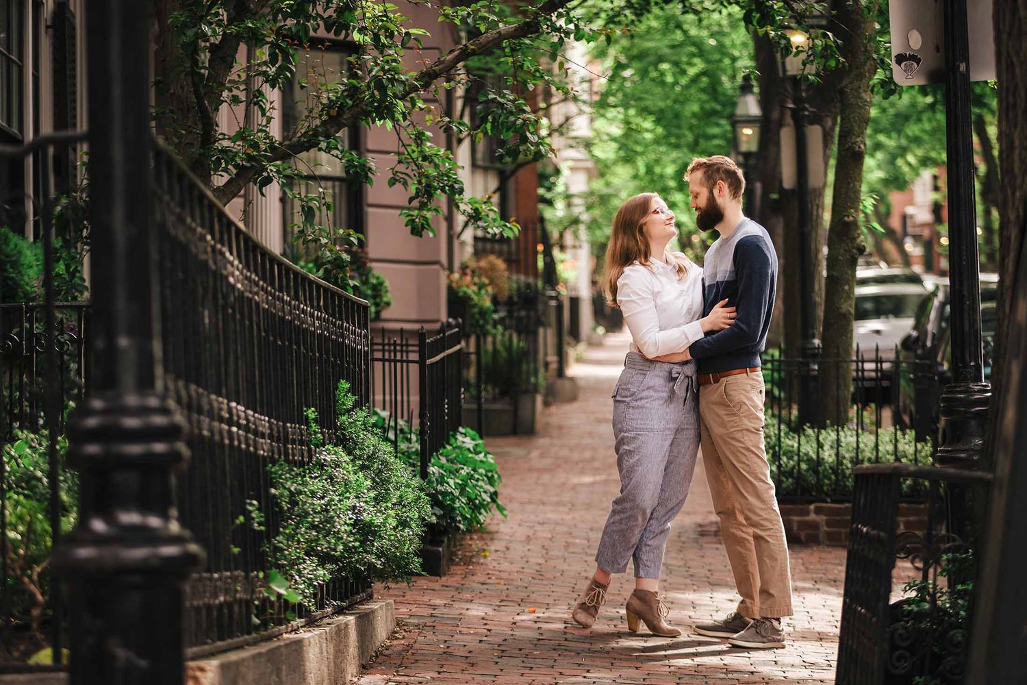 A wide-angle portrait of a couple embracing on a brick-lined Beacon Hill sidewalk, framed by iron fences, lush greenery, and gaslit lanterns