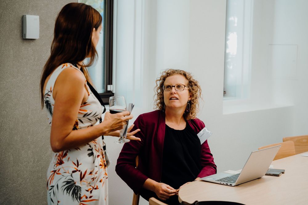 Two colleagues have a discussion at a desk with a laptop in a bright office setting.
