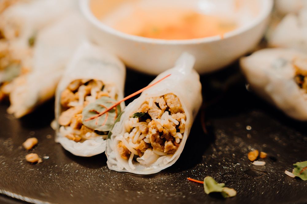 Fresh spring rolls filled with vegetables and rice noodles served with orange-colored dipping sauce on dark slate.