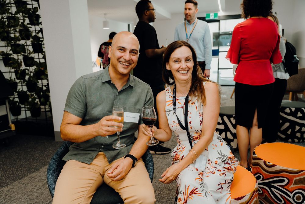 Two people in casual attire sit together enjoying wine at a social gathering with others visible in background.