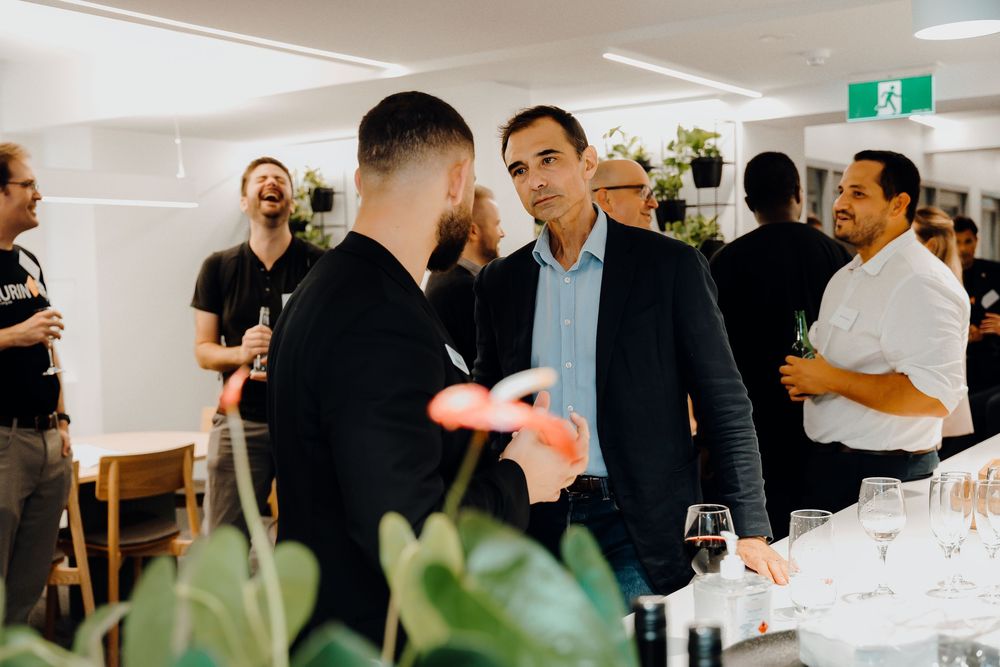 Two people in business attire engaged in conversation at an upscale social gathering with wine glasses and guests visible.
