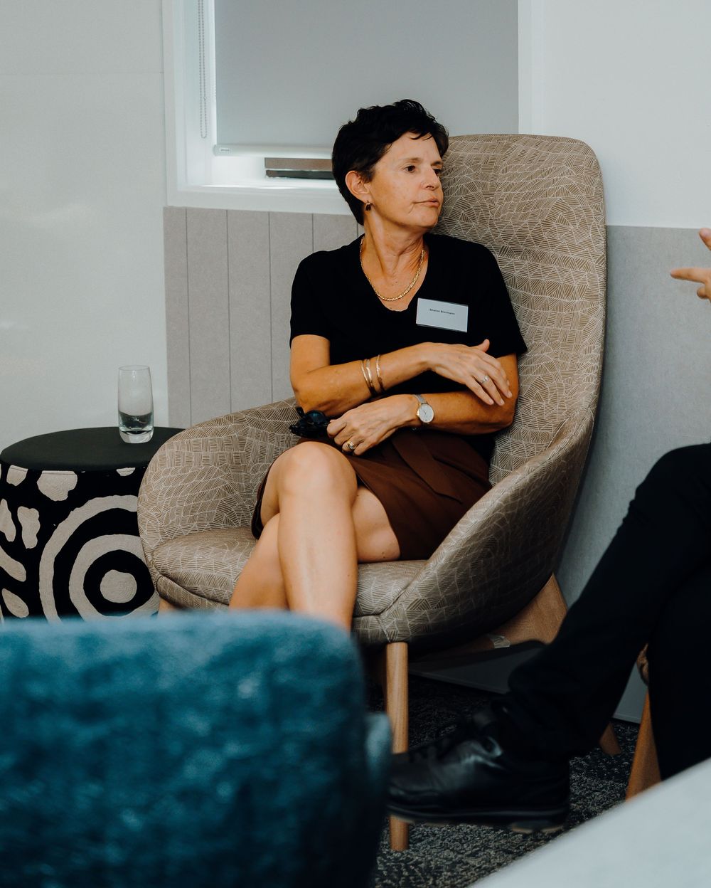 Professional wearing a black shirt sits contemplatively in a beige armchair during what appears to be a business meeting.