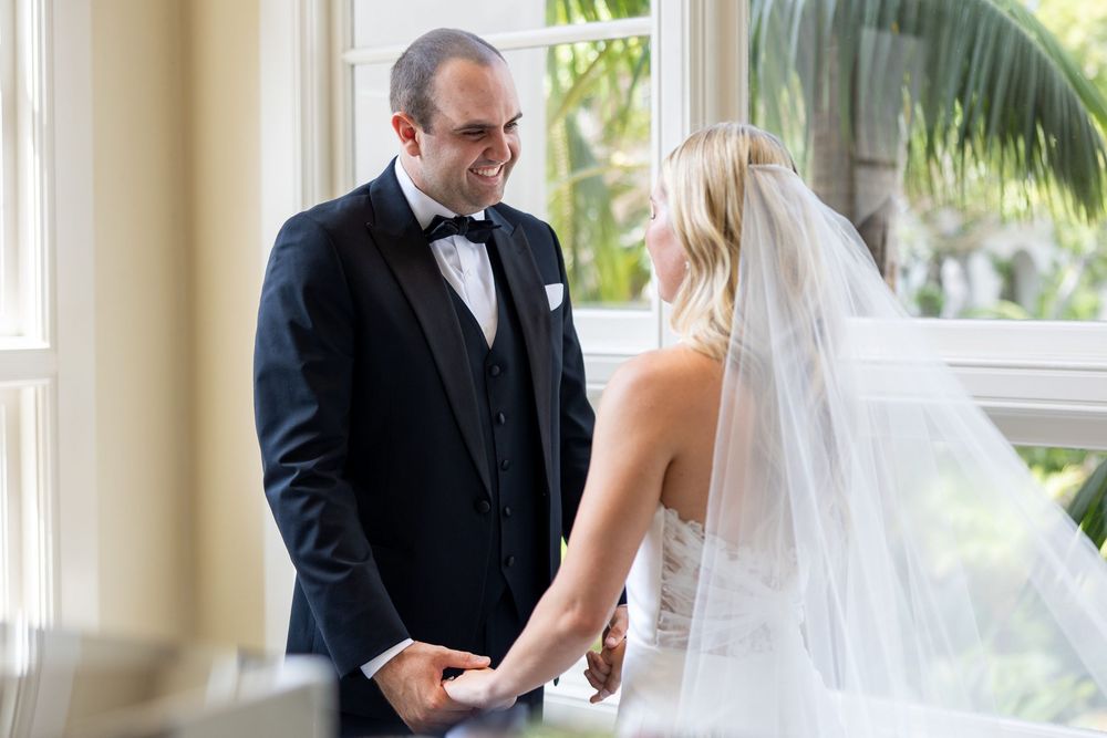 A couple exchanges rings during an elegant indoor wedding ceremony with sunlit windows and tropical greenery in view.