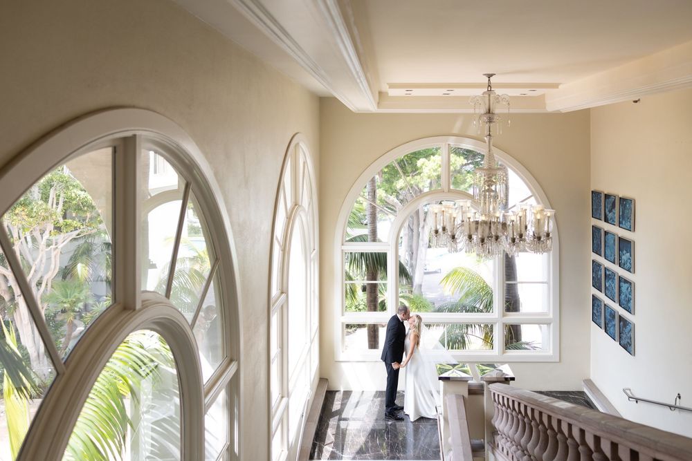 Curved archways extend down a beige hallway with palm trees visible through windows in an elegant building interior.