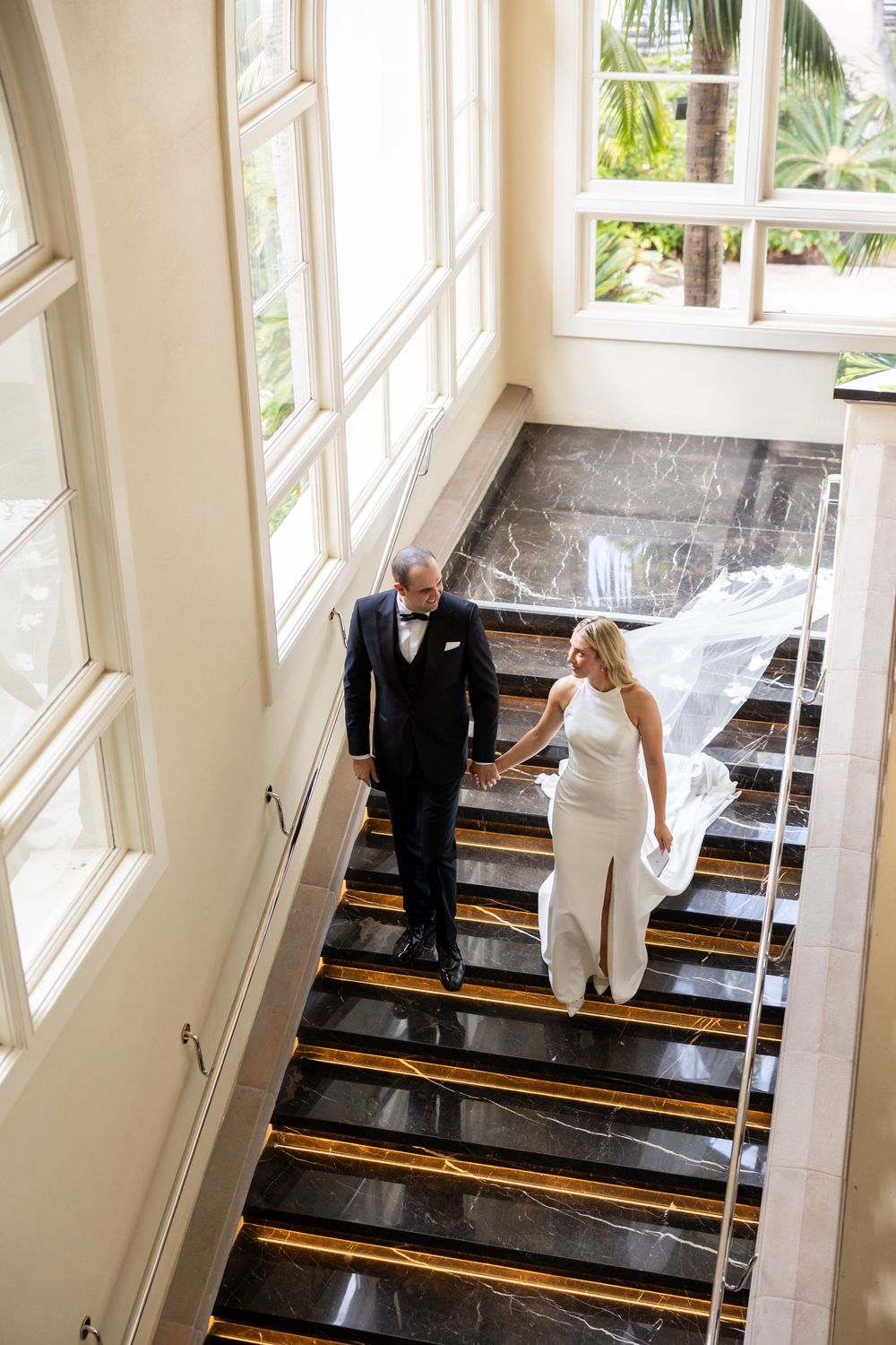 A couple stands together on an elegant black marble staircase with large windows letting in natural light.