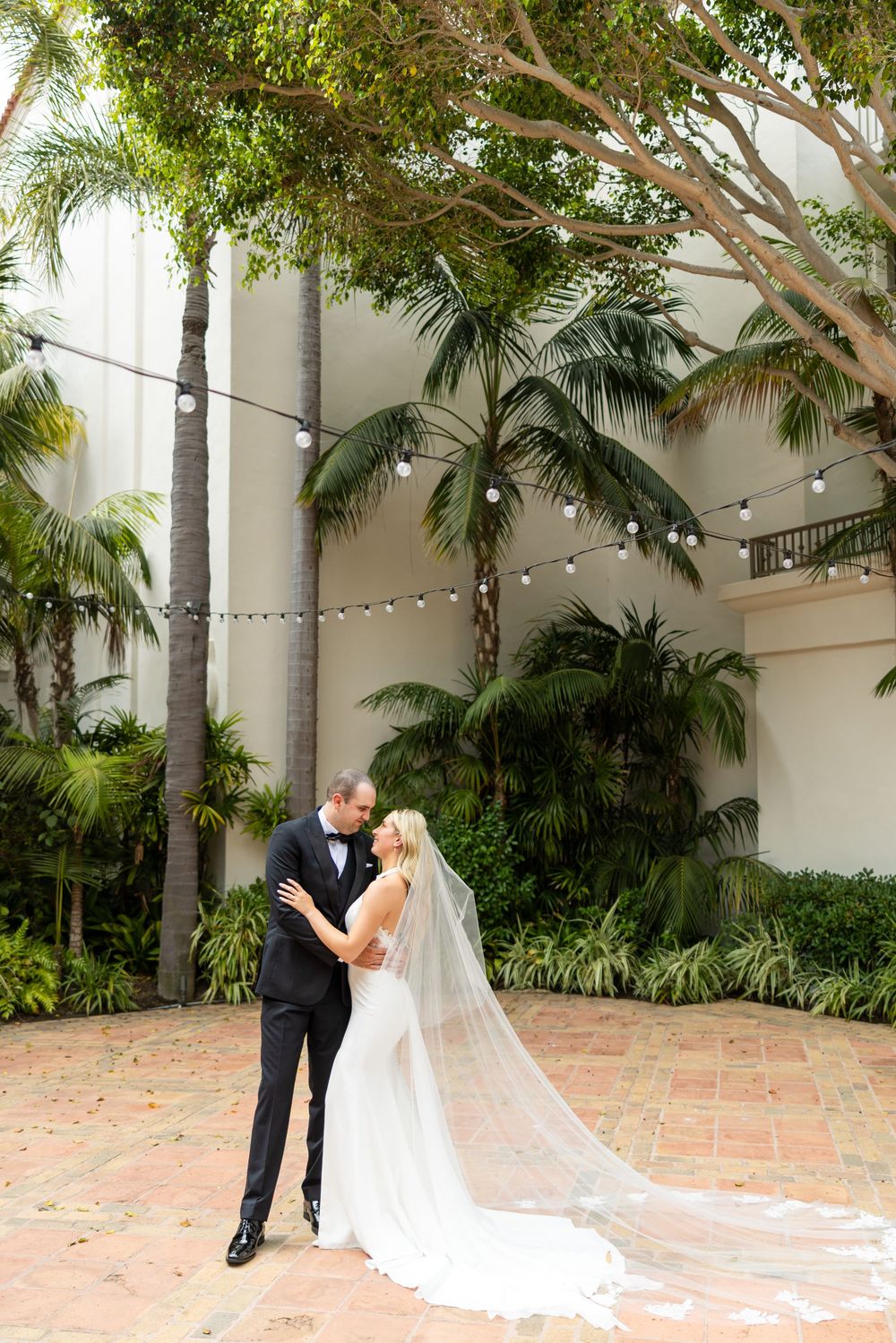 Two people share an intimate moment surrounded by tropical palm trees and string lights in an elegant courtyard setting.