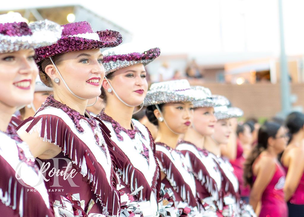 A row of cheerleaders in maroon uniforms and sparkly silver hats stand together at an outdoor event.