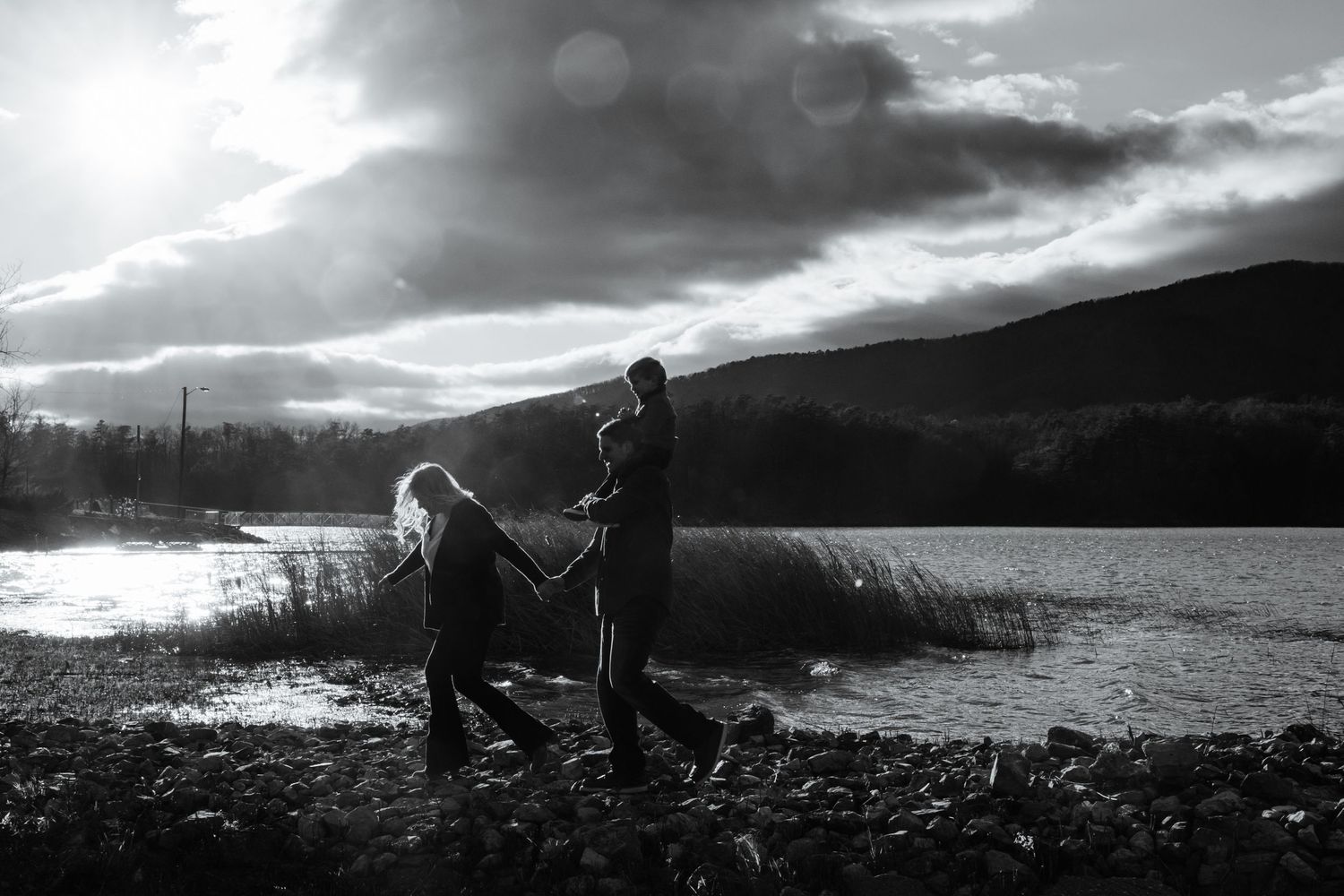 Black & white photo of a family walking hand-in-hand along a lakeside at sunset, with dramatic skies and a cild riding on shoulders