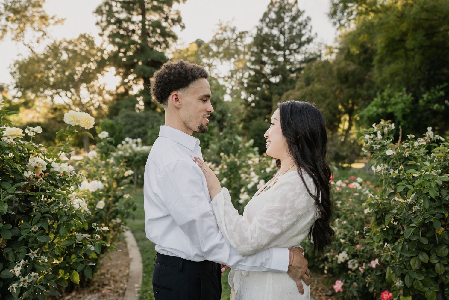 A couple shares a romantic moment in a blooming rose garden at sunset, wearing matching white outfits.
