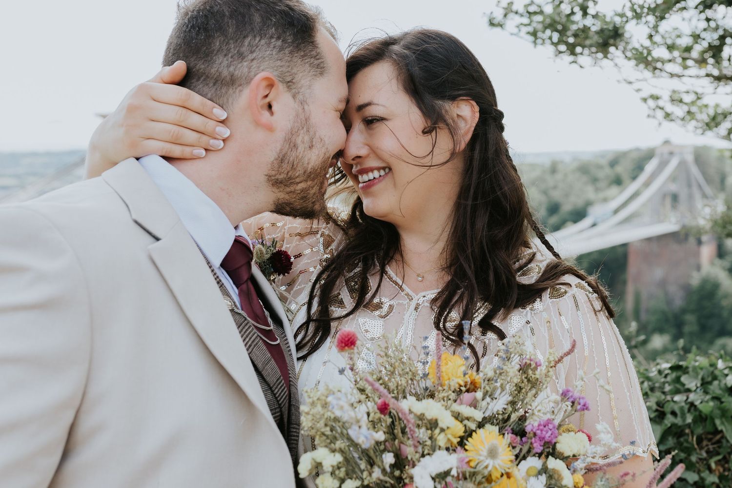 A romantic wedding moment captures a loving couple embracing outdoors with a wildflower bouquet and natural scenery.