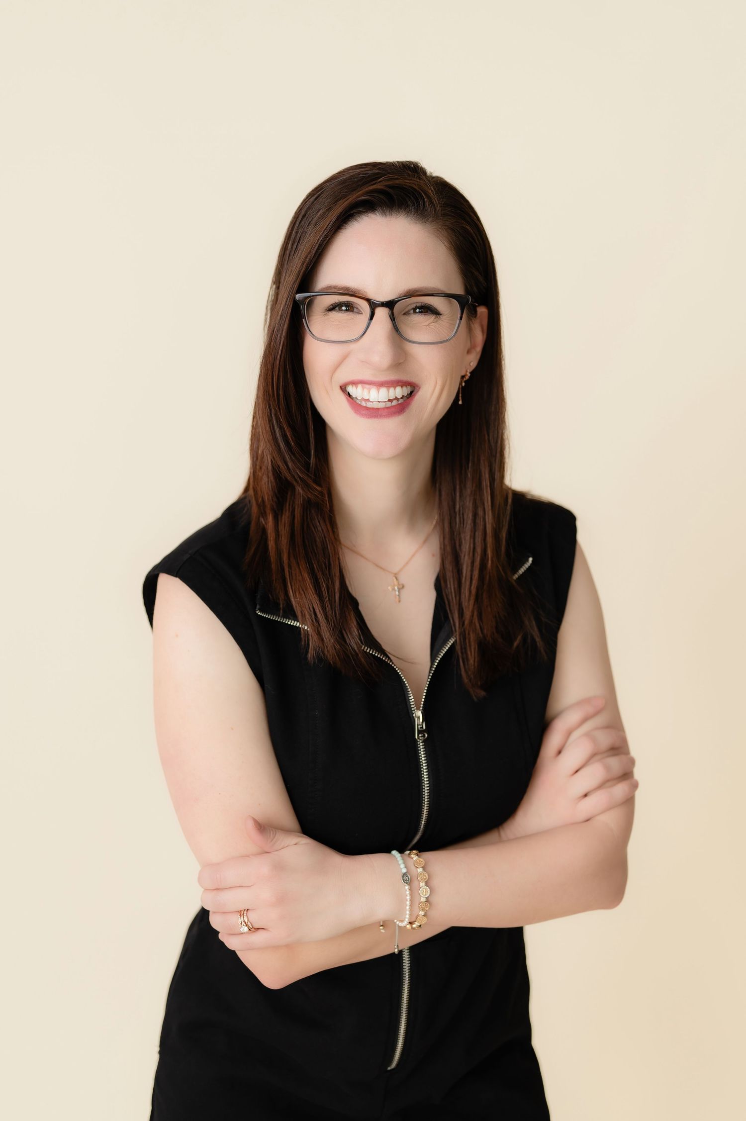 Professional headshot of a person wearing glasses and a black sleeveless top against a beige background, smiling warmly.