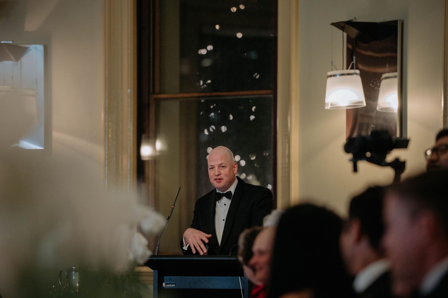 A speaker stands at a podium during an elegant evening event with soft lighting and formal decor.